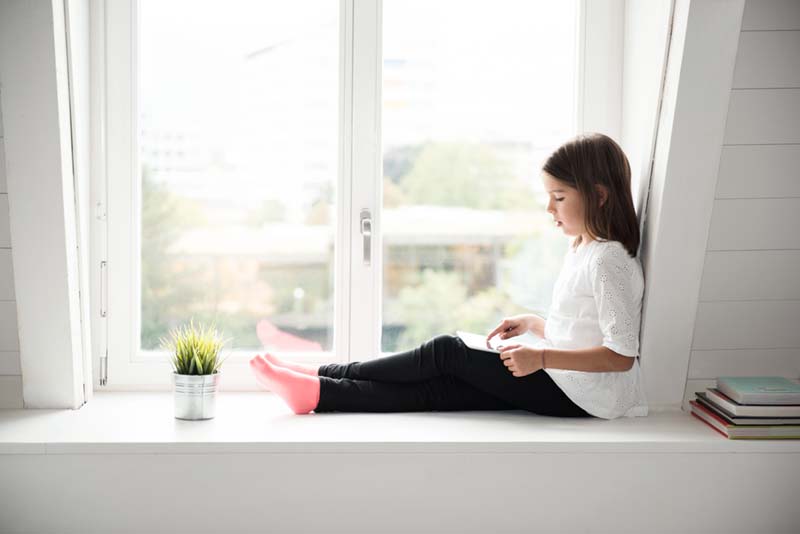 Tyttö lukee ikkunalaudalla e-kirjaa